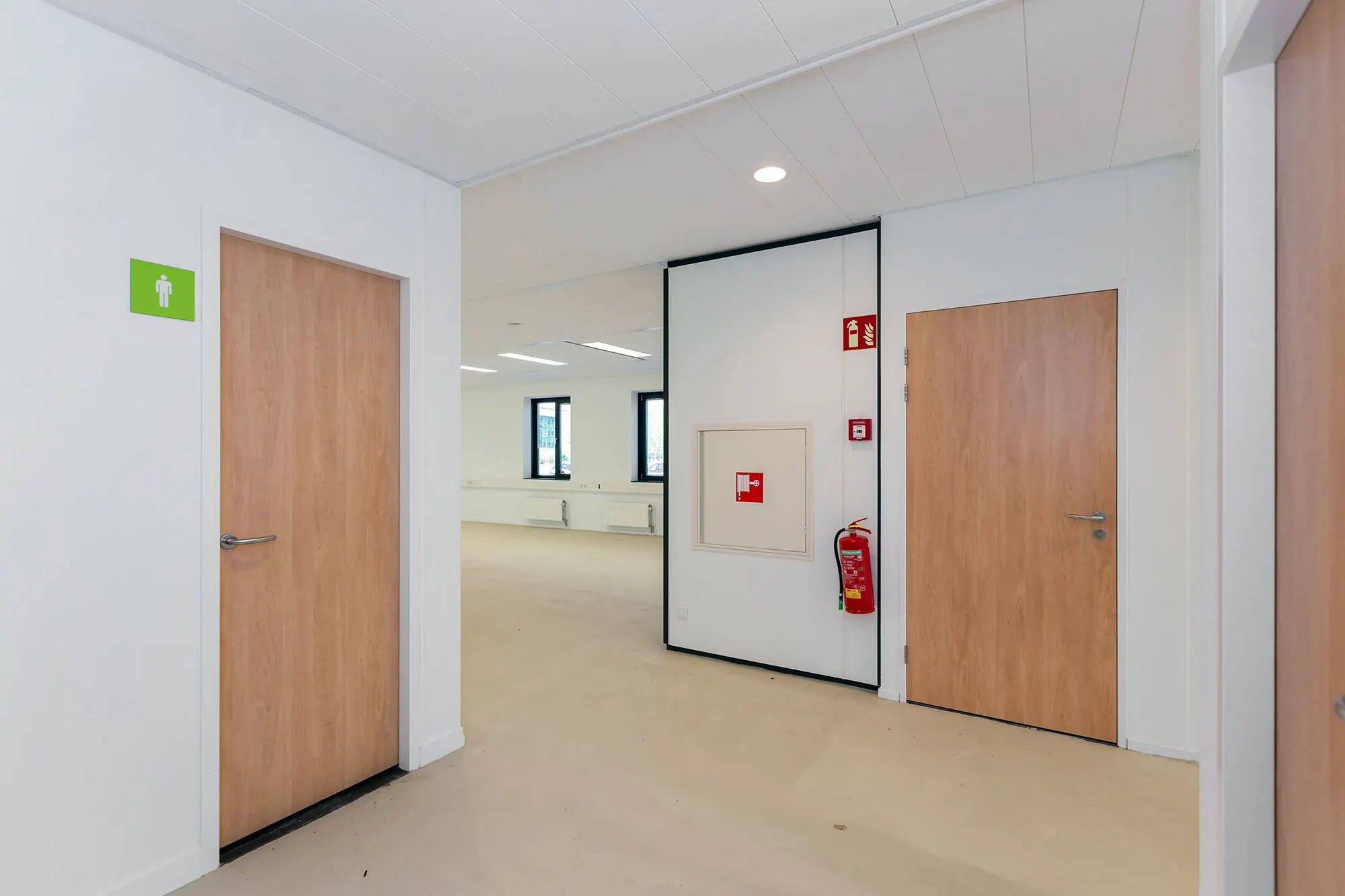 Hal met deuren, waaronder een toiletdeur met groen pictogram, en een brandslanghaspel met brandblusser aan de muur in een kantoorpand aan de Boris Pasternaklaan.